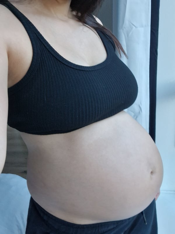 A pregnant woman standing sideways in a tank top, showcasing her belly near a window.