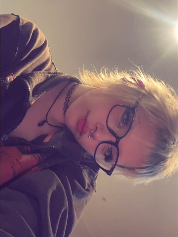 A young woman with blonde hair and glasses wearing a choker necklace, smiling softly in casual clothing.