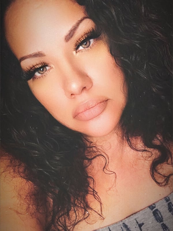 A woman with long curly dark hair and bold makeup posing confidently in a close-up selfie.