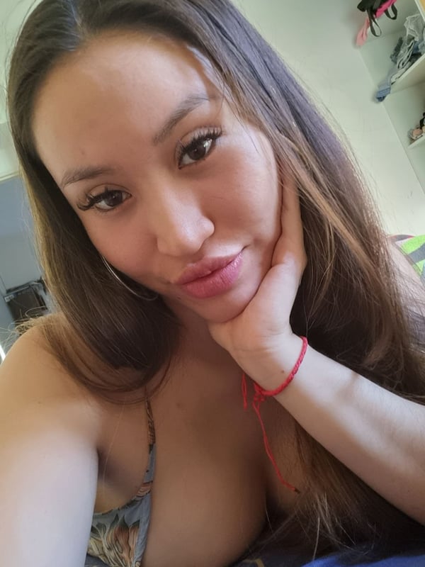 A woman taking a selfie in a low-cut top, smiling with hand on cheek in a home setting.