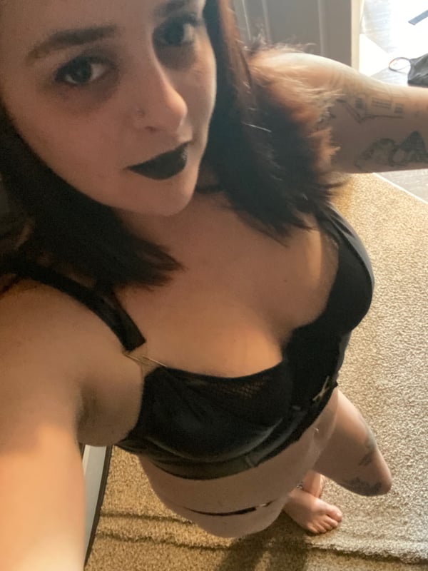 Close-up portrait of a woman with brunette hair, dark lipstick, and tattoos, posing confidently indoors wearing a black top.