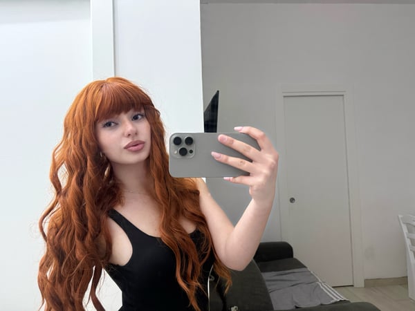 A woman with long red hair posing on a bed in red lingerie, looking back over her shoulder.