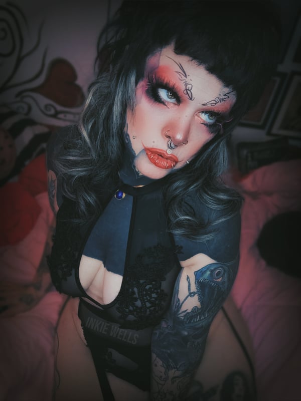 A close-up portrait of a woman with face tattoos, piercings, and dramatic makeup, wearing a black top with a keyhole neckline, against a dimly lit background.