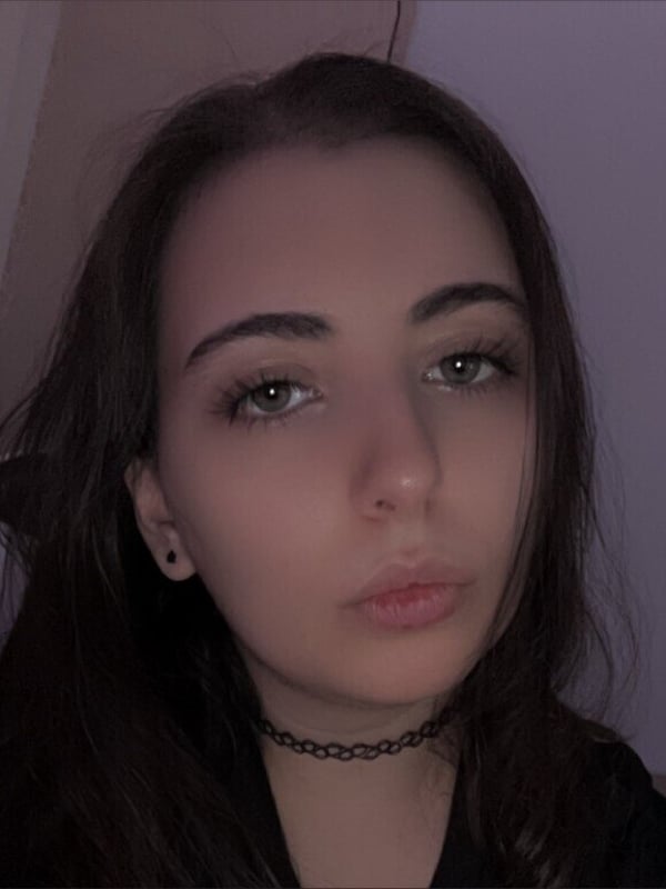 A close-up portrait of a young woman with long dark hair, wearing a black choker and small earrings, looking directly at the camera.