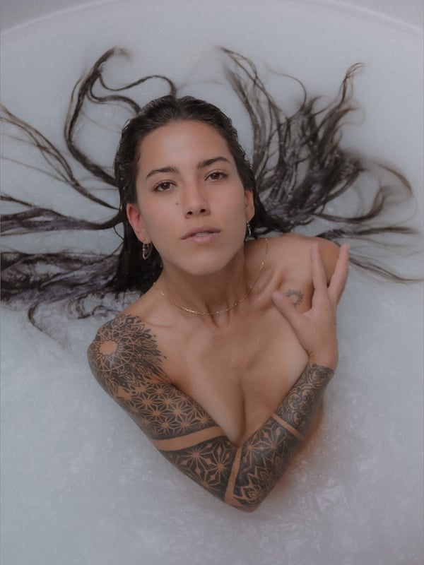 A woman relaxing in a bathtub filled with bubbles, with wet hair and tattoos visible on her arms.