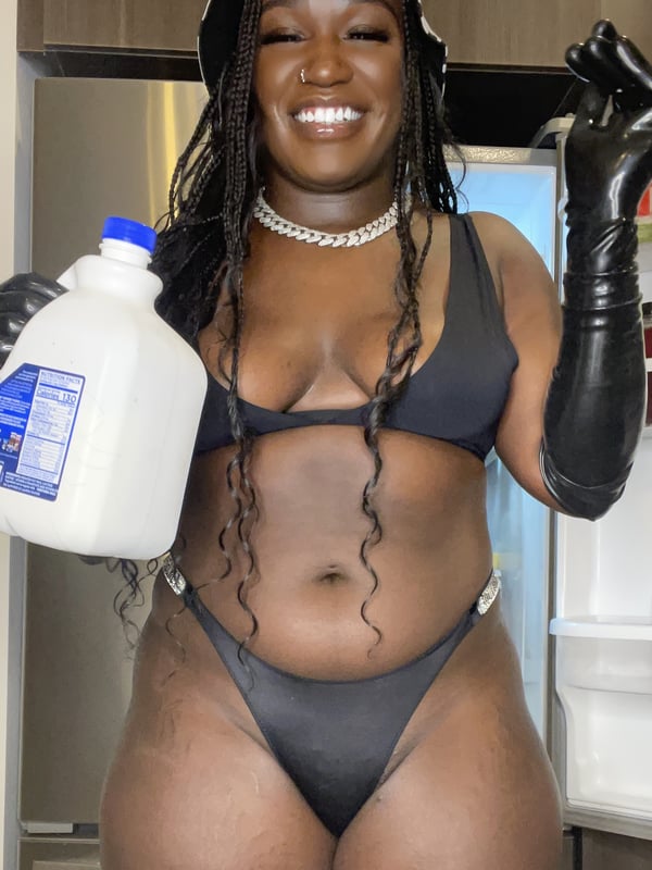 A smiling woman with braided hair wearing a bucket hat, black bikini, and long black gloves, holding a white milk jug in a kitchen setting.
