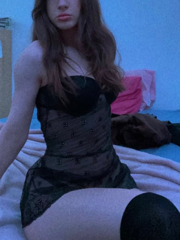 A young woman with long wavy hair sitting on a bed in a blue-lit room, dressed in a sheer black top.