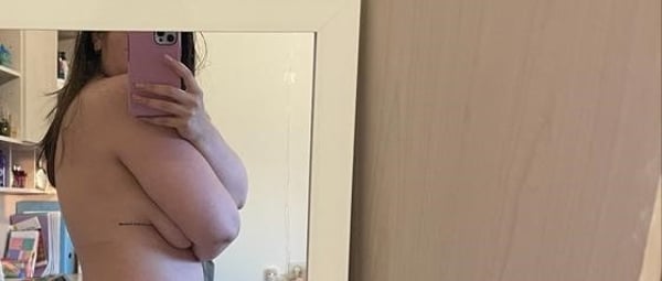 A young woman stands in front of a mirror in a bedroom, holding her phone and smiling softly with arms crossed over her chest.