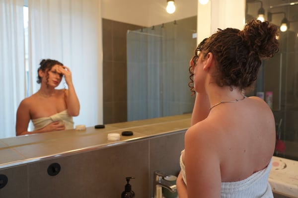A woman with curly hair wrapped in a towel stands in a modern bathroom, looking at her reflection in the mirror.