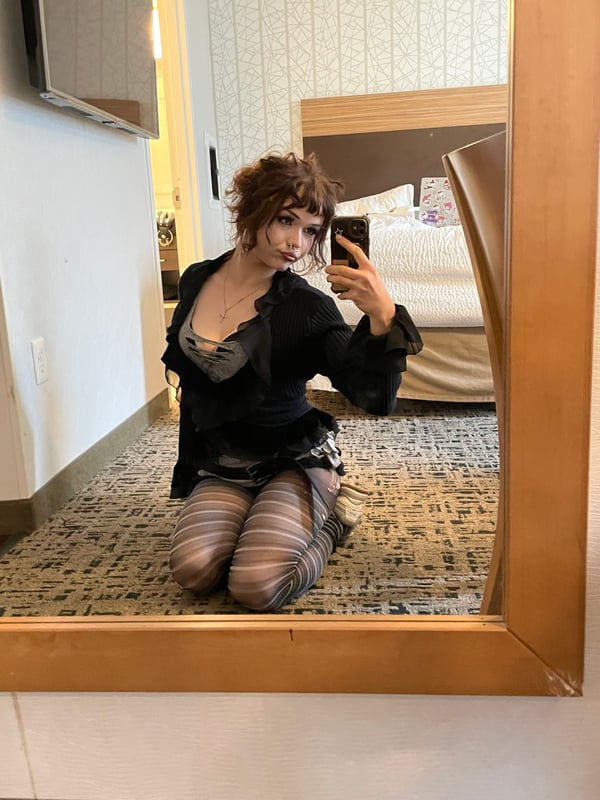 Selfie of a person with brown hair and bold makeup wearing a black lace top and a graphic jacket.