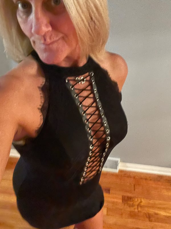 A mature woman with blonde hair in a close-up selfie, wearing a black lace-up top, standing indoors with a wooden floor.