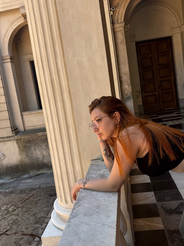 Young woman with red hair leaning against a stone balustrade in an architectural outdoor setting.
