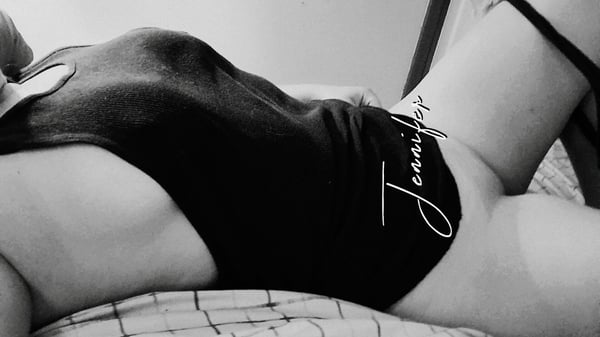 A woman reclining on a bed in a form-fitting black outfit, posing confidently with tattoos visible.