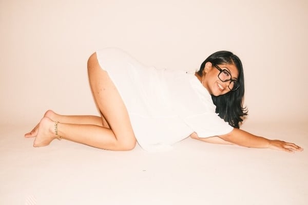Young woman with glasses and long dark hair posing playfully on the floor in a white t-shirt against a light background.