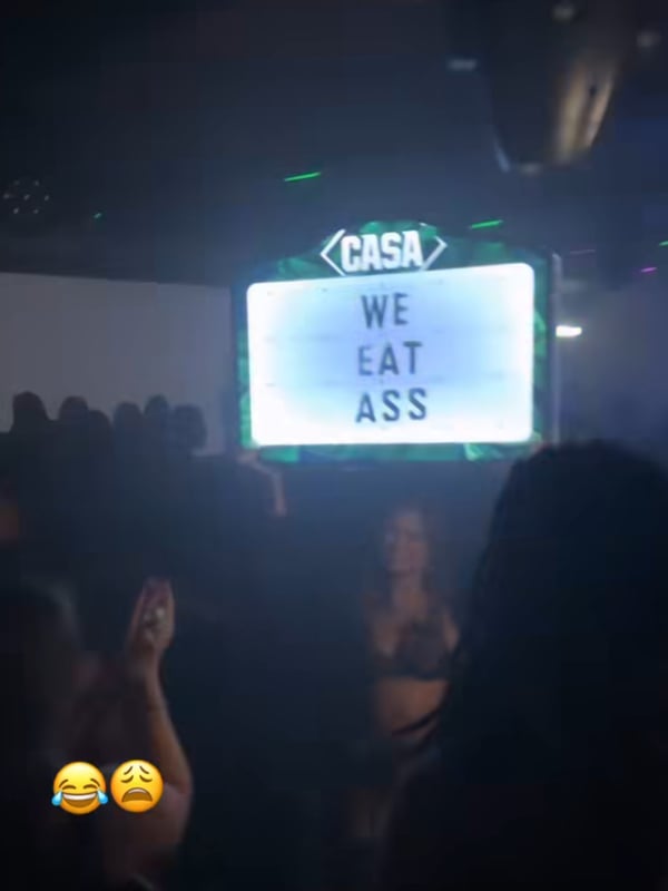 A woman posing in a revealing outfit at a vibrant nightclub with a neon sign reading 'We Eat Ass' in the background.