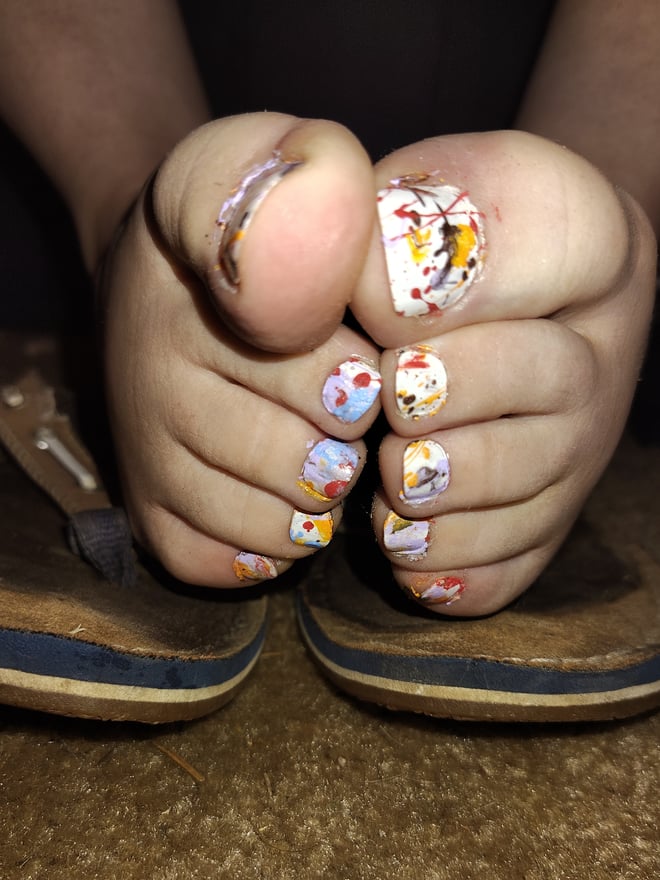 Close-up of bare feet with multicolored painted toenails positioned in front of worn brown flip-flops.
