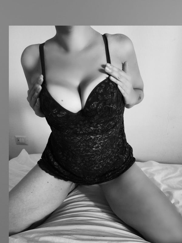 A woman sitting on a bed in a black lace top, posing confidently in a black and white photo.