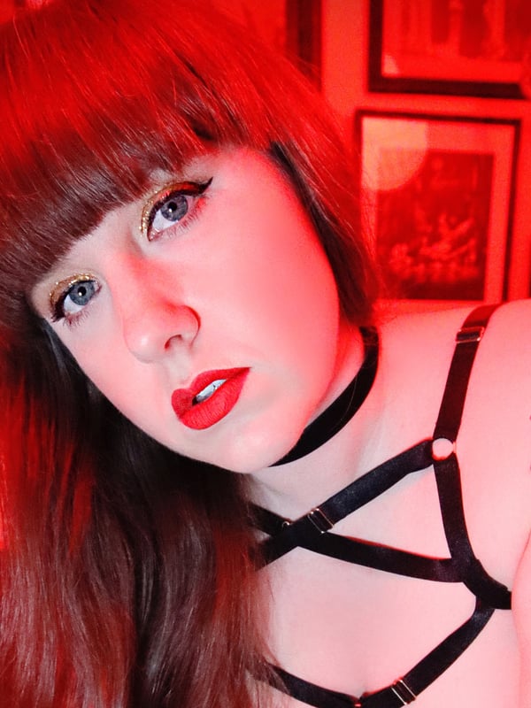 A woman with tattoos and bold makeup poses in front of framed artwork in a red-lit room.