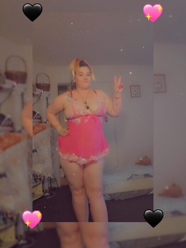 Person posing playfully in pink lingerie with peace sign gesture, featuring dyed blonde hair and tattoos, in front of a mirror.