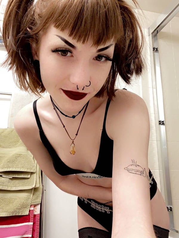 Young woman with short brown hair in pigtails, bold makeup, and a choker necklace posing in front of a bathroom mirror.