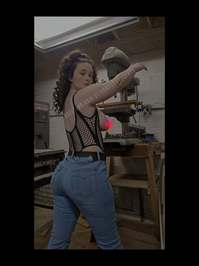 A young woman with curly hair posing confidently in a workshop setting, wearing a black top and jeans near industrial machinery.