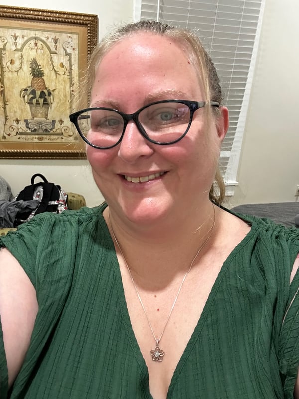 A smiling woman with glasses and blonde hair wearing a green top, standing indoors near a framed picture.