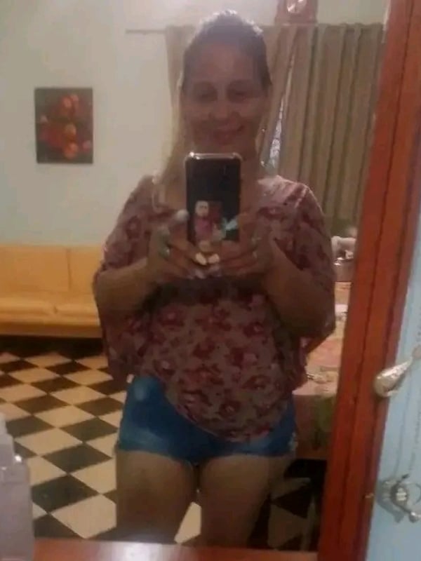 A smiling woman with loose dark hair holds her phone for a selfie in a room with checkered flooring, yellow sofa, and artwork on the wall.