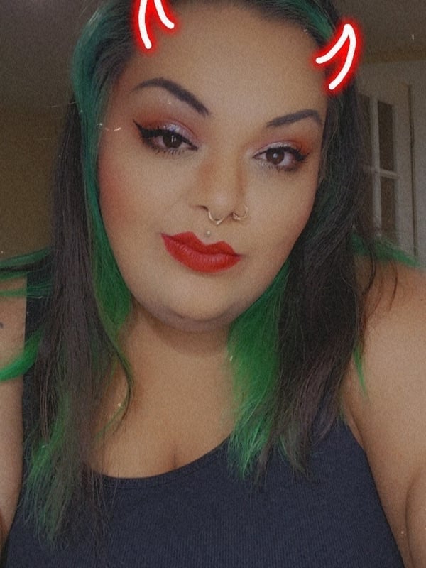 A woman with green hair and red devil horns smiling in a casual tank top indoors.