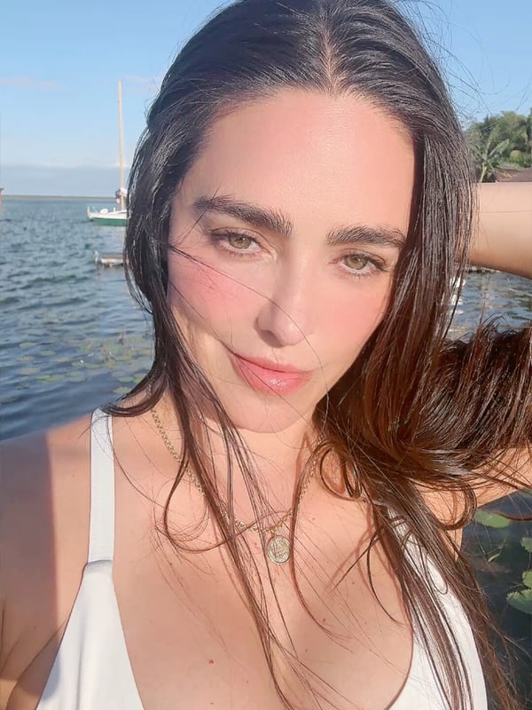 A woman smiling outdoors near water with a sailboat in the background, wearing a white swimsuit top.