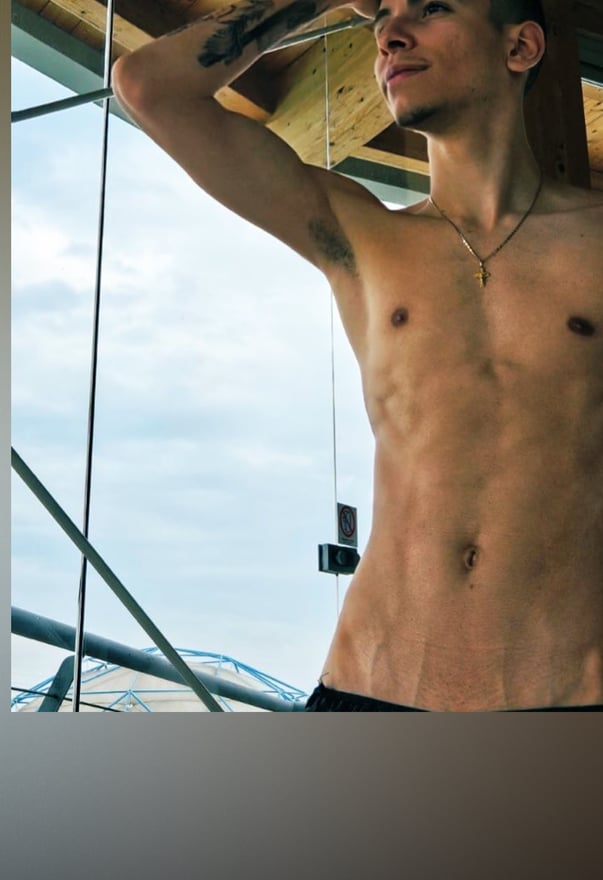 Young athletic man posing shirtless outdoors with arm raised, showing tattoos and muscular build against a construction-like background.