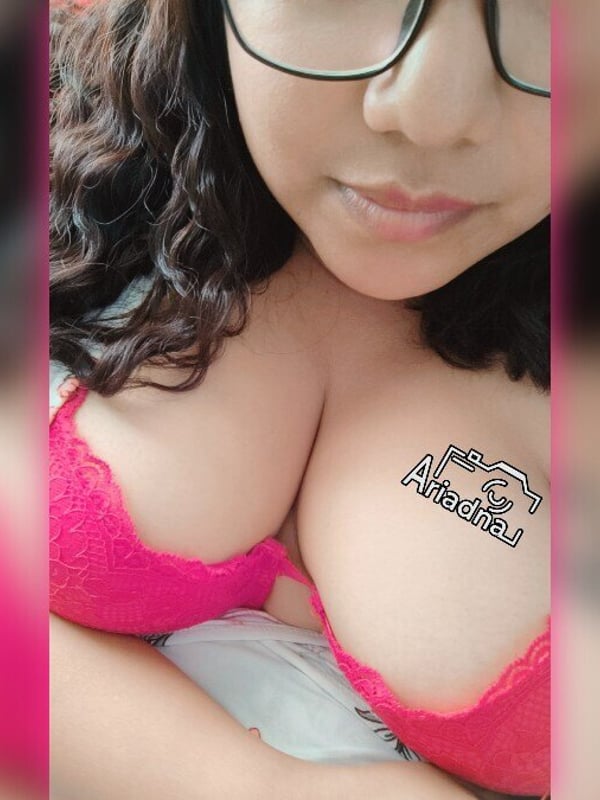 Woman with glasses and curly dark hair smiling at the camera, wearing pink lingerie.