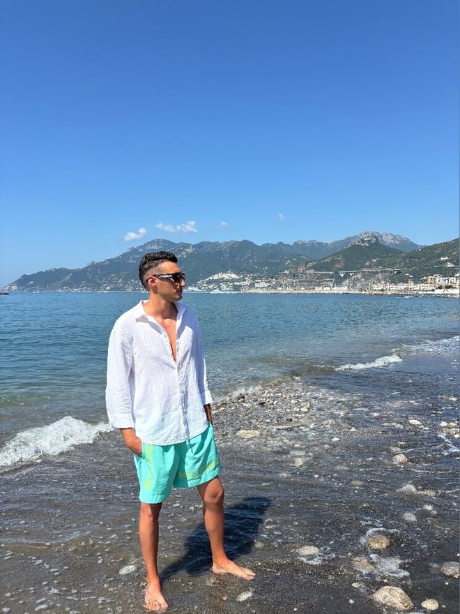 A man in a white shirt and green shorts stands on a rocky beach by the sea, wearing sunglasses and posing with mountains and blue water behind him.