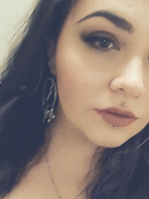 Close-up selfie of a young woman with dark hair, hoop earrings, lip makeup, and a lip piercing, smiling softly against a plain background.