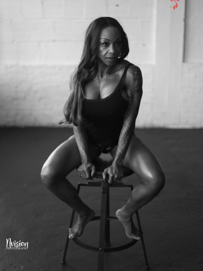 A woman with long hair posing confidently on a stool in a black tank top against a white brick wall.