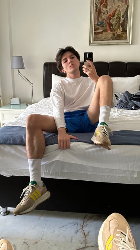 Young man lounging on a bed in a white long-sleeve shirt, blue shorts, and white sneakers with yellow accents, taking a mirror selfie in a bedroom with artwork on the wall.