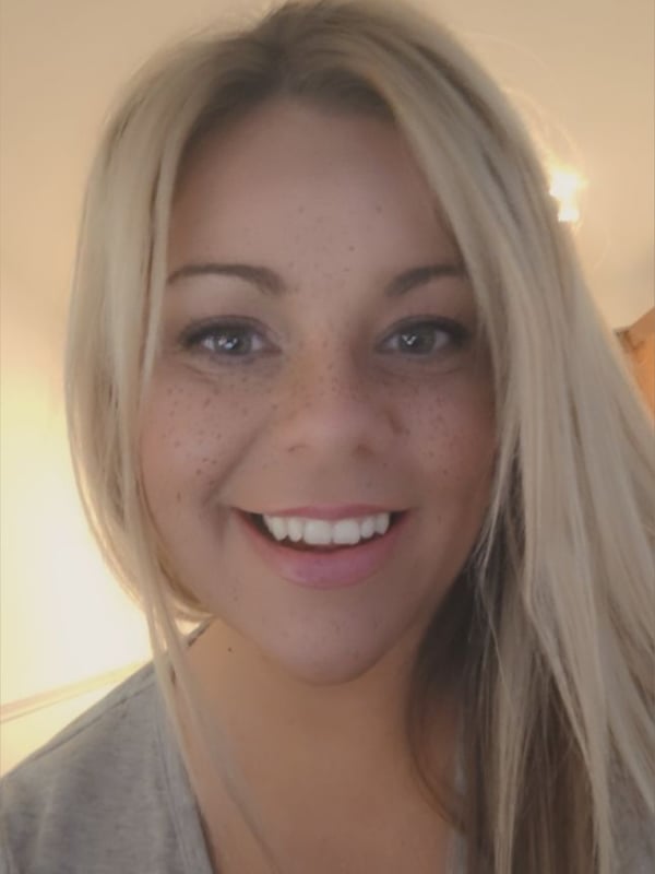 A smiling young woman with long blonde hair posing indoors with soft lighting.