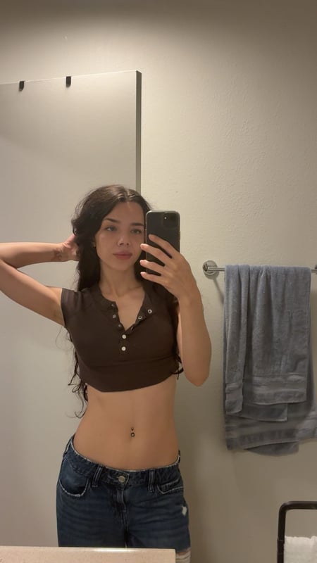 Young woman with long dark hair taking a mirror selfie in a black bikini top and patterned bottoms, standing in a bathroom.