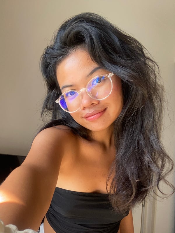 Young woman with long dark hair and clear glasses smiling in a casual black top, taking a selfie indoors.