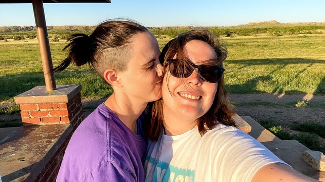 Two women smiling and kissing outdoors near a wooden structure in a grassy field under a sunny sky.