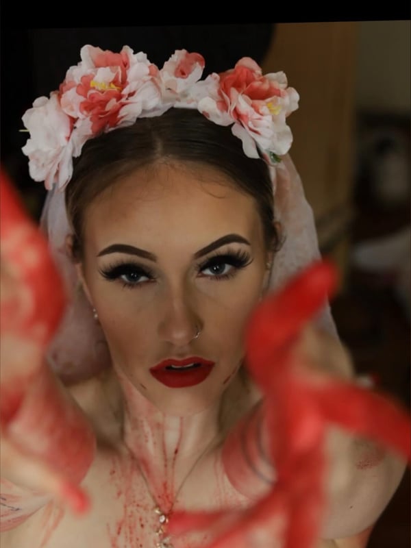 A woman with long blonde hair wearing a white veil adorned with pink and red flowers, posing with red-stained hands near her face in dramatic makeup.