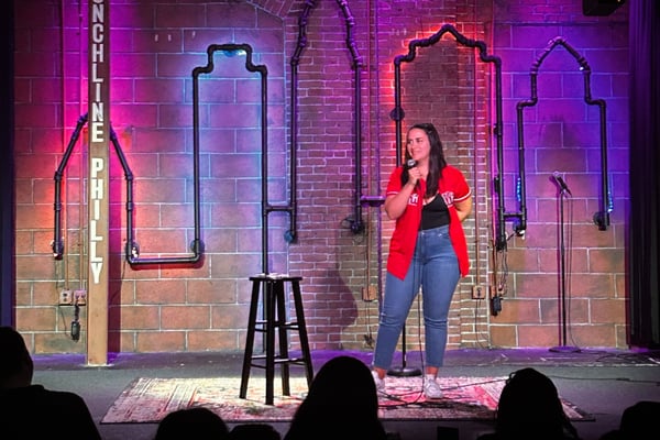 A woman performing stand-up comedy on stage in a red blazer and jeans, illuminated by colorful neon lights against a brick wall backdrop with a microphone stand.