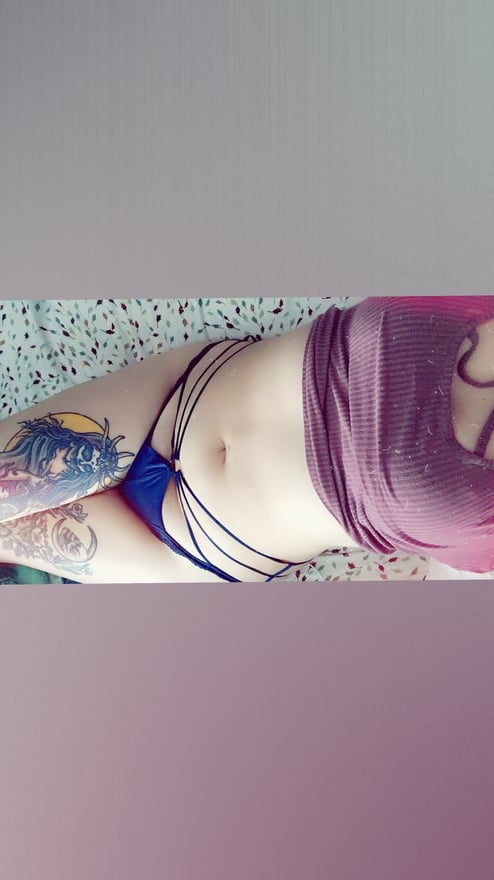 A woman lying on a surface wearing a purple top and strappy bottoms, with colorful tattoos visible on her skin.