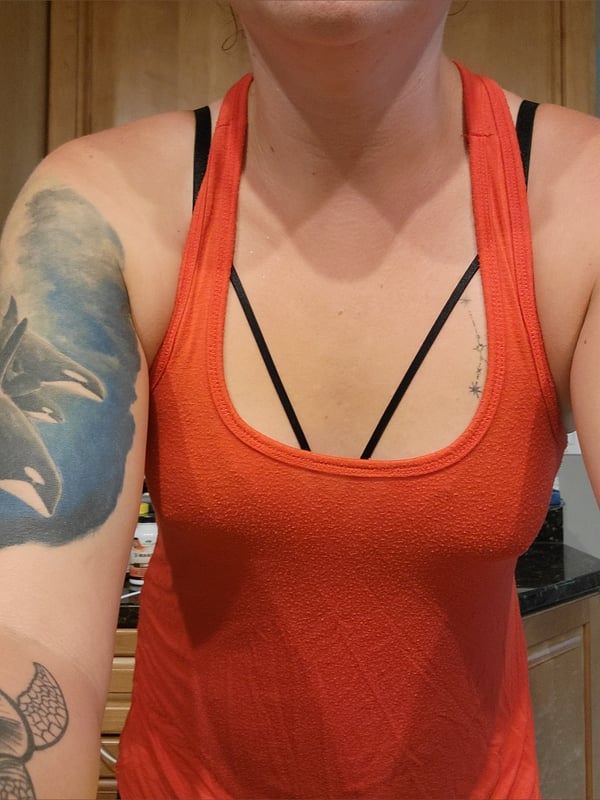 A woman standing in a kitchen wearing an orange tank top with black straps, displaying tattoos on her arm.