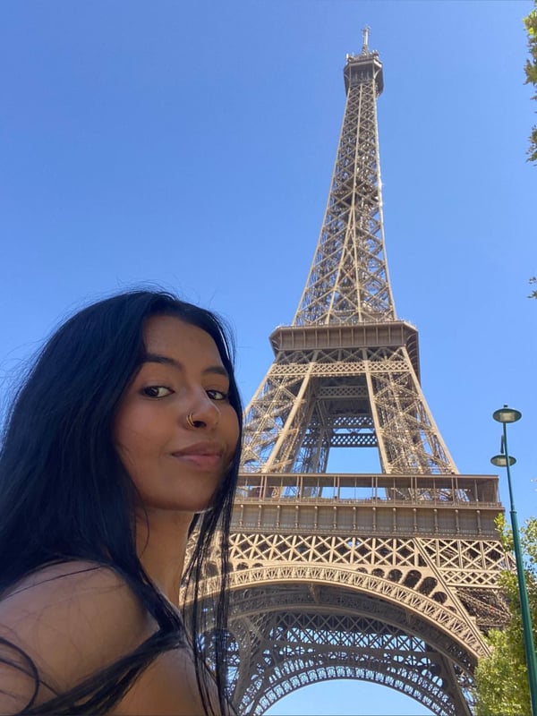 A young woman with long dark hair smiling in front of the Eiffel Tower under a clear blue sky.