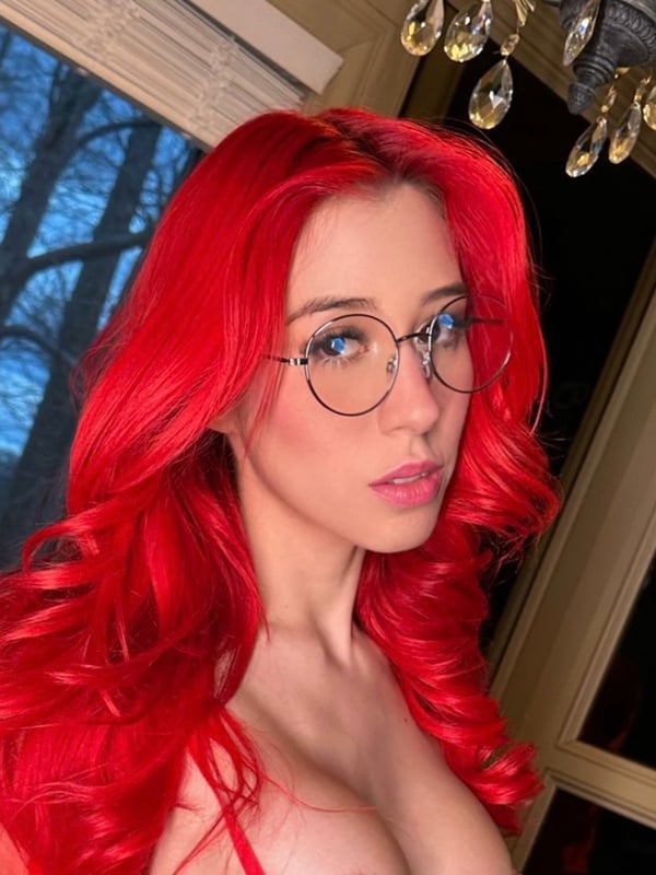 A woman with long red hair and glasses smiling softly indoors near a window with elegant chandelier lighting and a scenic outdoor view.