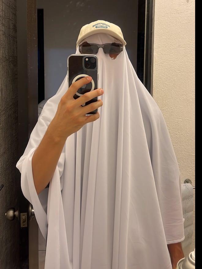A person wearing a white hooded garment, cap, and sunglasses, taking a selfie indoors near a door.