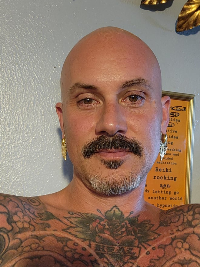 A bald man with a mustache, earrings, and tattoos smiling at the camera indoors with decorative background elements.
