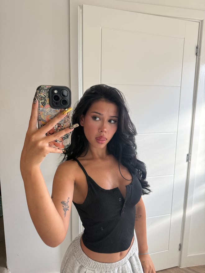 A young woman taking a selfie indoors while wearing a gingham top and holding a decorated phone case.