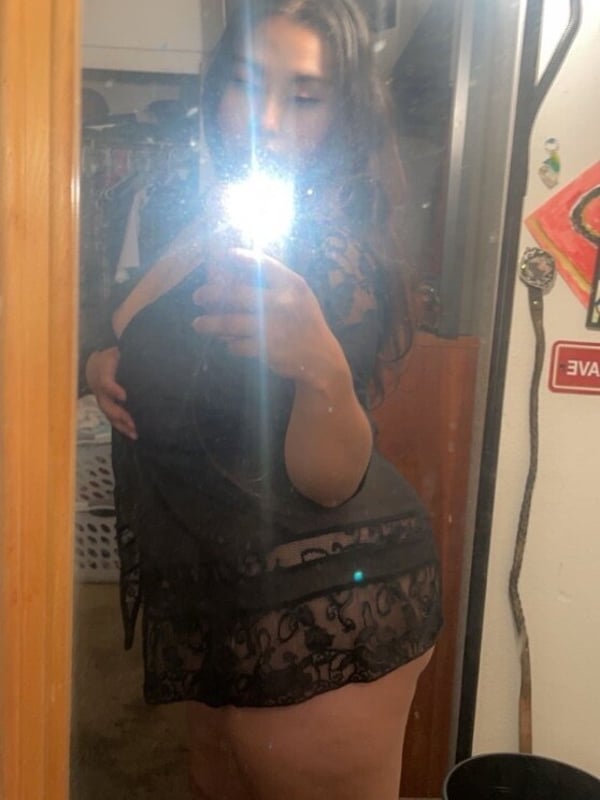 A woman posing in a lace outfit in front of a bathroom mirror, holding her phone for a selfie with soft lighting.