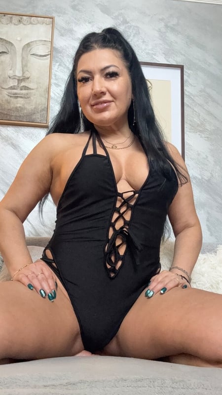 Woman with long dark hair posing confidently in a black swimsuit inside a room with a Buddha statue on the wall.
